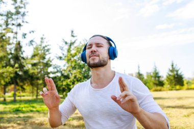 Mutlu adam kulaklıkla müzik dinliyor açık havada beyaz ceketli yaz parkında.