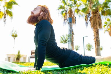 Uzun sakallı yakışıklı kızıl saçlı adam güneşli sahilde yoga yaparken siyah spor takım elbise giyiyor..