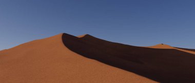 Arkasında büyük bir tepe olan bir çöl manzarası. Gökyüzü mavi ve güneş parlıyor.