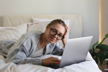 Evde, yatağında gözlük ve süveter takan ve dizüstü bilgisayarla çalışan esmer bir kadının portresi. Uzaktan kumandalı iş. Arkaplan, kopyala boşluk, kapat.