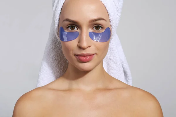 Young woman wearing a towel after showering, wearing eye patch for dark circles treatment. Female applying cosmetic products to decrease puffiness under her eyes. Close up, background.