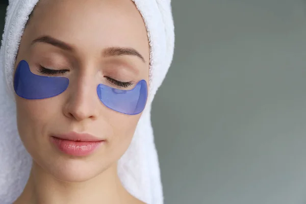 Young woman wearing a towel after showering, wearing eye patch for dark circles treatment. Female applying cosmetic products to decrease puffiness under her eyes. Close up, background.