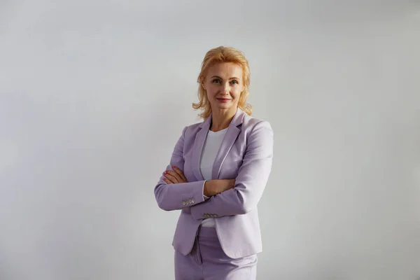 Portrait of beautiful blonde businesswoman standing with arms crossed over the chest. Confident adult woman wearing lilac jacket standing with folded hands. White wall background, copy space, close up