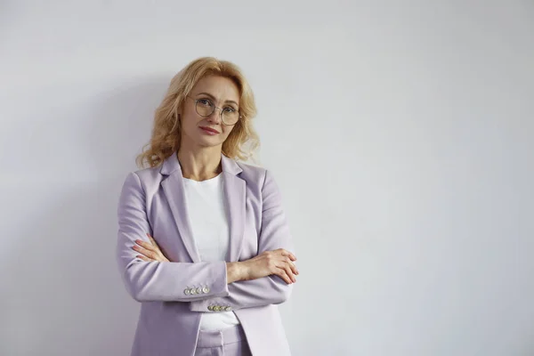 Portrait of beautiful blonde businesswoman standing with arms crossed over the chest. Confident adult woman wearing lilac jacket standing with folded hands. White wall background, copy space, close up