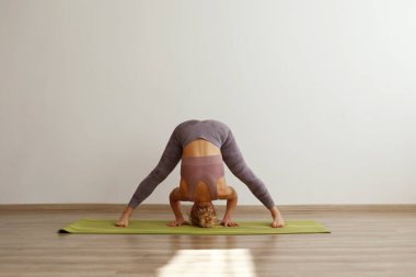Sporty adult woman practicing hatha yoga at home. Fit middle aged yogini doing the wide legged forward bend pose. White wall background, copy space, close up.