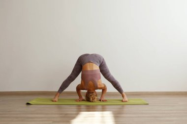 Sporty adult woman practicing hatha yoga at home. Fit middle aged yogini doing the wide legged forward bend pose. White wall background, copy space, close up. clipart