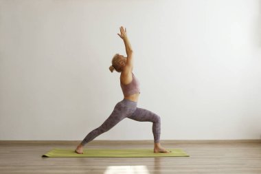 Sporty adult woman practicing hatha yoga at home. Fit middle aged yogini doing the warrior I pose. White wall background, copy space, close up.
