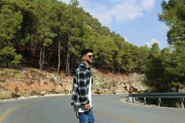 Young man walking in the middle of a switchback road alone. Hipster guy wearing checked flannel shirt and sunglasses wandering outdoors. Copy space, coniferous forest background.