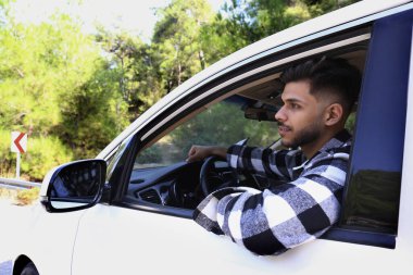 Young carefree man wearing a checked flannel shirt looking out of the window of white car. Smiling guy enjoying his road trip. Close up, copy space, background.