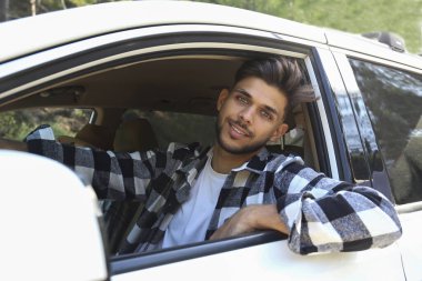 Young carefree man wearing a checked flannel shirt looking out of the window of white car. Smiling guy enjoying his road trip. Close up, copy space, background.