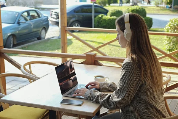 Young beautiful woman with wireless headphones working remotely from coffee shop patio. Female freelancer typing on her laptop. Close up, copy space, background.
