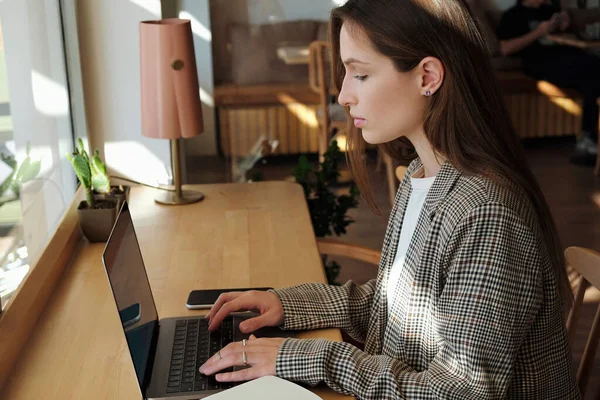 Beautiful young woman sitting in coffee shop with laptop and a planner. Female freelancer wearing smart casual outfit working in coffeehouse. Remote job concept. Close up, copy space, background.