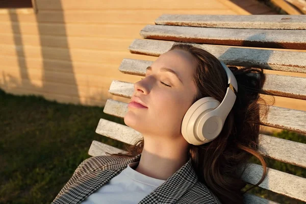 一个年轻美丽的女人的画像 她的面部表情轻松地躺在木制的甲板椅子上 听着她最喜欢的音乐 柔和的日落光 复制空间 — 图库照片