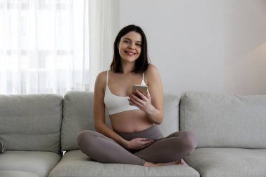Pregnancy tracking app concept. Young pregnant woman sitting on the couch at home and having a video call on the phone. Close up, copy space, background.