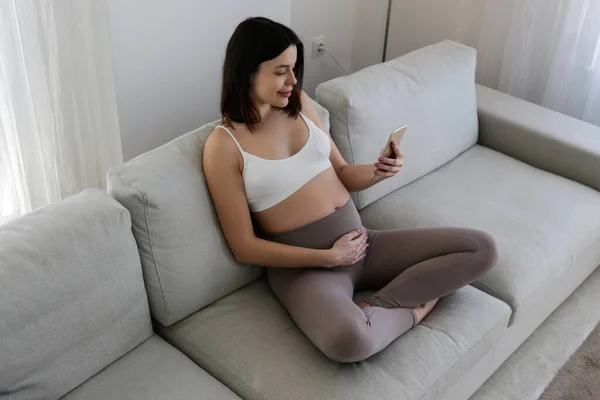 Pregnancy tracking app concept. Young pregnant woman sitting on the couch at home and having a video call on the phone. Close up, copy space, background.