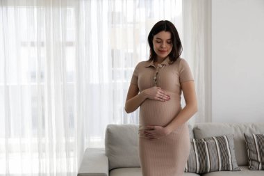 Young beautiful woman on second trimester of pregnancy. Pregnant female wearing maternity wear, with arms around her belly. Expecting a child. Background, copy space