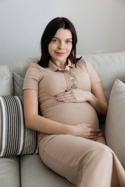 Young beautiful woman on second trimester of pregnancy. Pregnant female wearing maternity wear, with arms around her belly. Expecting a child. Background, copy space