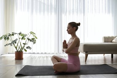 Young woman practicing hatha yoga at home. Yogini wearing colorful sportswear sitting in namaste pose with her palms pressed together in front of the chest. Copy space, background.