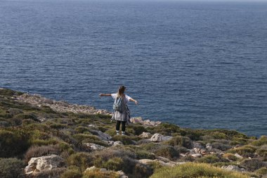 Atlantik Okyanusu 'nun kayalık kıyı şeridinde yürüyüş yapan genç bir kadının arka görüntüsü. Yeşil tepede deniz tarafından yıkanmış bir kadın yürüyüşçü. Yalnız seyahat konsepti. Boşluğu kopyala, arkaplan.
