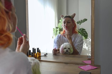 Çok renkli saçlı genç bir kadın aynanın karşısında oturuyor ve kucağında bir köpeğe dudak kremi sürüyor. Sabah güzellik rutini konsepti. Kapat, kopyala boşluk, arkaplan.