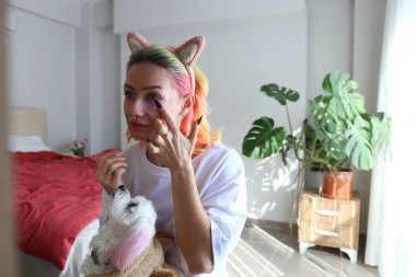 Çok renkli saçlı genç bir kadın aynanın karşısında oturuyor ve göz bantları altında kucağında bir malta köpeğiyle oturuyor. Sabah güzellik rutini konsepti. Kapat, kopyala boşluk, arkaplan.