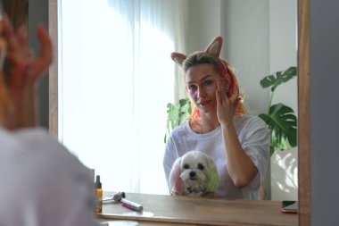 Çok renkli saçlı genç bir kadın aynanın karşısında oturuyor ve göz bantları altında kucağında bir malta köpeğiyle oturuyor. Sabah güzellik rutini konsepti. Kapat, kopyala boşluk, arkaplan.