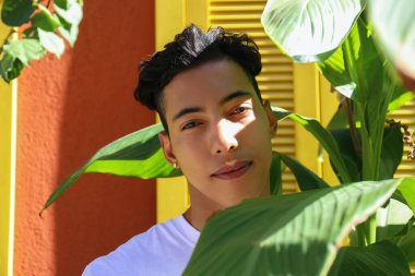 Kırmızı duvarlı ve sarı pencereli kırsal evin yakınındaki palmiye yapraklarının arasında duran düz beyaz tişörtlü yakışıklı Latin adam. Kapat, kopyala boşluk, arkaplan.