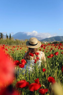 Çevre dostu bir çiçek çiftliğinde kırmızı haşhaş tarlasının ortasında beyaz pamuklu elbise ve hasır şapka giyen genç bir kadın. Kapat, boşluğu kopyala, arkaplanda dağlar.