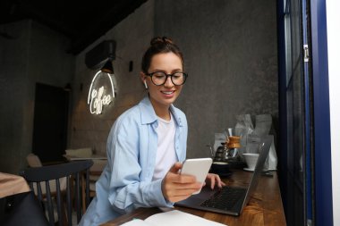 Bir kafede dizüstü bilgisayarıyla oturmuş telefonuna bakan şık giyinmiş güzel bir kadının portresi. Uzaktan çalışma konsepti. Kapat, kopyala boşluk, arkaplan.