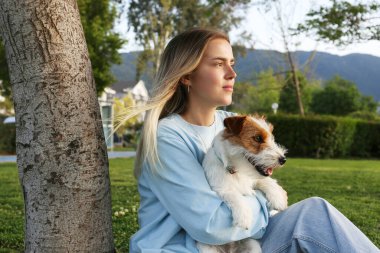 Genç bir kadın yeşil çimlerde oturuyor ve tel saçlı Jack Russell Terrier köpeği ile bir ağaca yaslanıyor. Sarışın bir kadın ve parktaki kaba bir köpek. Boşluğu kopyala, arkaplan.