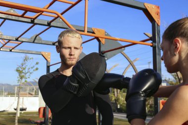 Kişisel antrenörüyle antrenman yapan ve açık hava spor salonunda pedlere vuran genç bir kadın. Temiz havada boks seansı. Kapat, kopyala boşluk, arkaplan.