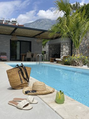 Yüzme havuzu temel konsepti. Güneş banyosu, güneş gözlüğü, hasır şapka ve buz gibi limonata için malzeme dolu bir plaj çantası. Köy evinin arka bahçesinde boş bir havuz. Boşluğu kopyala, dağ arkaplanı.