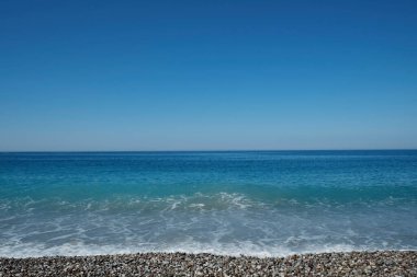 Güneşli bir günde bulutsuz açık mavi gökyüzü ile çakıl taşlı kumsalın güzel deniz manzarası. Ufuk çizgisi olan okyanus görünümü, metin için bir sürü kopya alanı olan arkaplan.