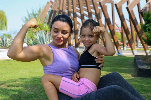 Super mom flexing her powerful bicep with her daughter. Sporty mother and her little girl are bonding through physical exercise. Close up, copy space, background.