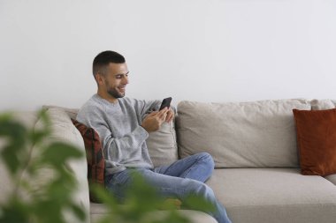 Köşedeki koltukta oturan sakallı adam. Elinde telefon var. Evde yalnız başına rahat bir adam. İç arkaplan, kopyalama alanı, kapat.