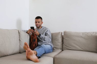 Köşedeki koltukta oturan sakallı adam. Elinde telefon var. Evde yalnız başına rahat bir adam. İç arkaplan, kopyalama alanı, kapat.