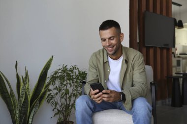 Koltukta oturan sakallı adam elinde telefonla gülümsüyor. Evde yalnız başına rahat bir adam. İç arkaplan, kopyalama alanı, kapat.