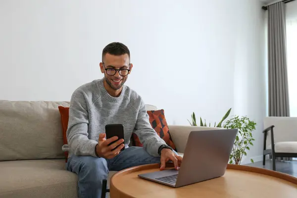 Happy young man wearing glasses looking at the screen of his phone with joy. Freelancer working on a couch in his living room. Close up, copy space, white wall background.