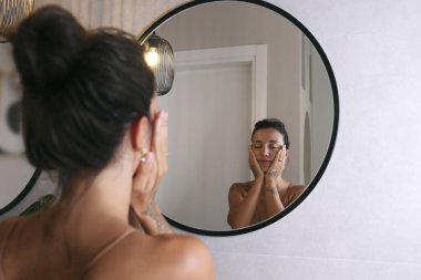 İpek gecelik giyen genç bir Orta Doğulu kadın aynaya bakıyor ve yüz masajı yapıyor. Sabah güzellik rutini konsepti. Kapat, kopyala boşluk, arkaplan.