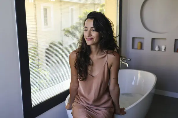 Young beautiful woman with brunette hair about to take a bath. Brunette female wearing silk nightgown sitting on the edge of the bathtub. Close up, copy space, background.