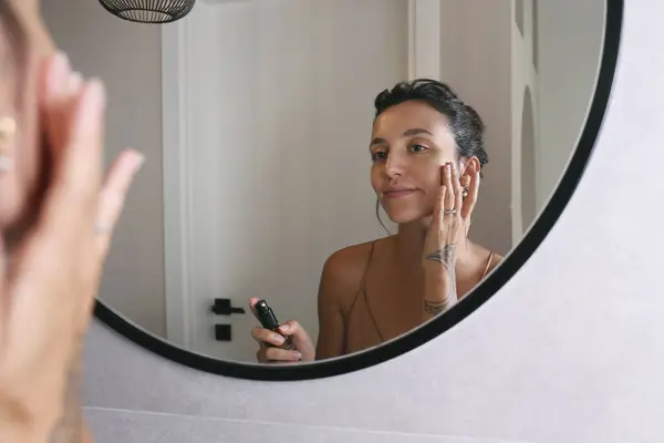 Young woman wearing a nightgown looking at the round shaped mirror and applying anti-aging skincare to her face. Morning beauty routine concept. Close up, copy space, background.