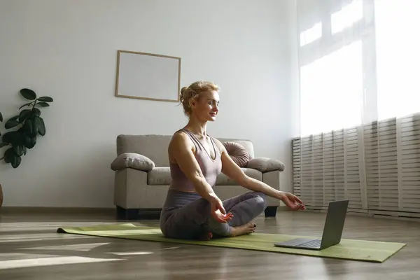 Sporty adult woman guiding a meditation in an online class. Fit middle aged yogini live streaming yoga masterclass. Interior background, copy space, close up.