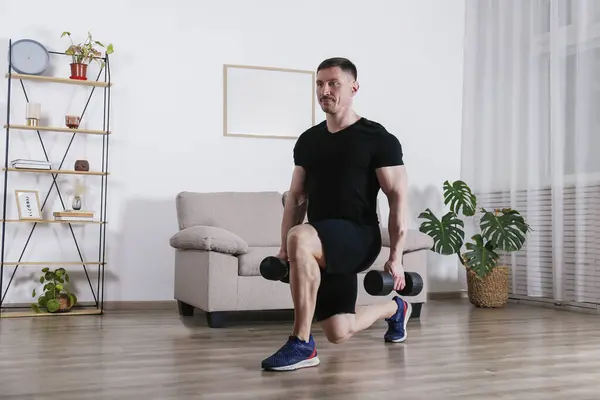 Bodybuilder training at home, doing lunges with dumbbells. Young man performing physical exercises in his living room. Close up, copy space, background.