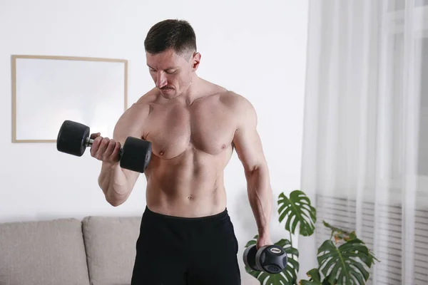 Bodybuilder training at home, doing supinated bicep curls with dumbbells. Young man performing physical exercises in his living room. Close up, copy space, background.