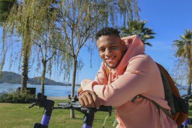 Kiralık bir e-scooter 'ın gidonuna yaslanan ve gülümseyen genç bir İspanyol. Parktaki motorlu mutlu latin adam. Kapat, kopyala alanı, yeşillik arkaplanı.