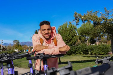 Kiralık bir e-scooter 'ın gidonuna yaslanan ve gülümseyen genç bir İspanyol. Parktaki motorlu mutlu latin adam. Kapat, kopyala alanı, yeşillik arkaplanı.