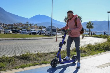 Güzel ada manzaralı mavi bir bisiklet yolunda e-scooter süren genç bir latin. Motorlu scooter 'la şehirden işe gidip geliyor. Boşluğu kopyala, tropikal hedef arkaplan.