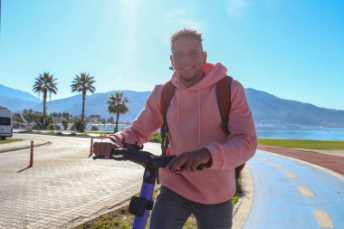 Güzel ada manzaralı mavi bir bisiklet yolunda e-scooter süren genç bir latin. Motorlu scooter 'la şehirden işe gidip geliyor. Boşluğu kopyala, tropikal hedef arkaplan.