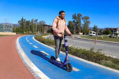 Genç Latin bir adam mavi bisiklet yolunda bir e-scooterla parktan çıkıyor. Motorlu scooter 'la şehirden işe gidip geliyor. Boşluğu kopyala, arkaplan.
