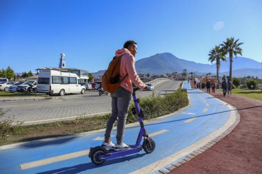 Güzel ada manzaralı mavi bir bisiklet yolunda e-scooter süren genç bir latin. Motorlu scooter 'la şehirden işe gidip geliyor. Boşluğu kopyala, tropikal hedef arkaplan.